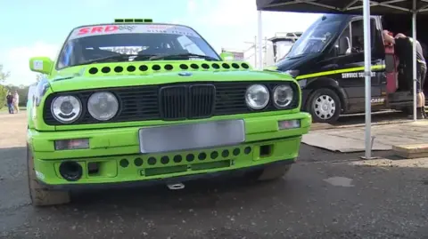 BBC A bright green rally car next to a black van with a blue sky in the background