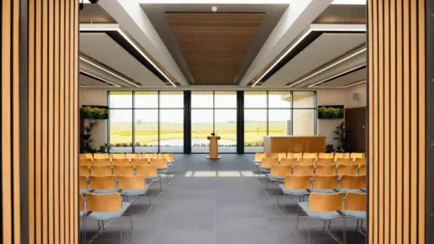 East Riding of Yorkshire Council A room with large floor to ceiling windows looking out onto a pond and grass. There is a grey tiled floor, five rows of seats on two sides separated by a space in the middle with a lectern at in front of the windows. There is a wood effect and white ceiling. There are two yellow wooden that can be drawn to close off the room. There are two photos of flowers on the wall and two to four floor standing plants in the distance. There is also a counter / bar area near the windows.