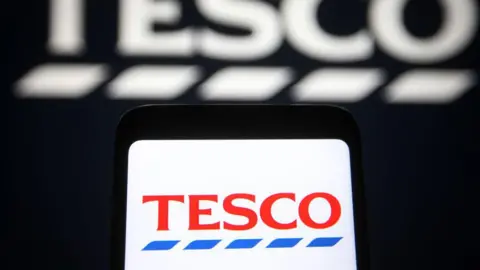 The Tesco logo is displayed on a smartphone, against a black background displaying a larger, blurred version of the supermarket's logo.