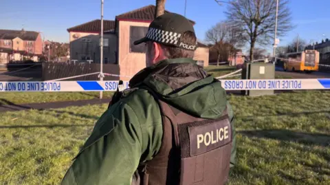 A police officer is standing with his back to the camera. It's daylight. He's standing next to a police cordon. There's a tape cordoning off the area.