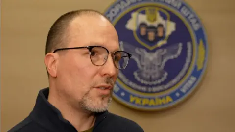 A man wearing glasses in front of the emblem of the Ukrainian military.