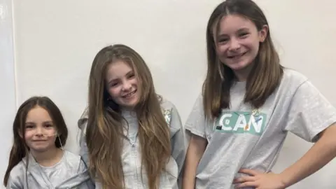 The three sisters are looking to camera in grey tops and smiling.