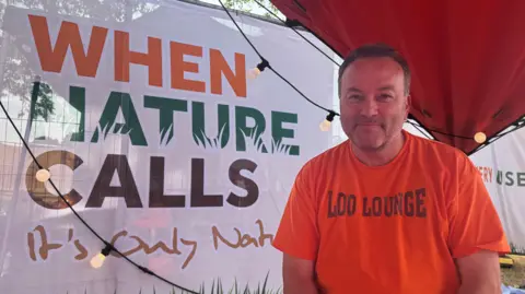 George King/BBC A head and shoulders image of a man wearing an orange T-shirt with Loo Lounge written on it. Next to him is a sign that reads When Nature Calls. 