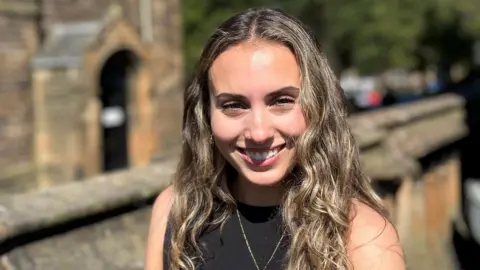 La BBC Orla Sonvico sourit à la caméra. Elle a de longs cheveux bruns et porte un haut noir. Au loin se trouve les murs et les vieux bâtiments de grès.