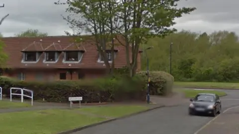 A Google Streetview image of a road with a car driving along it. There are houses on the side of the road and it is bordered with grass verges and hedges.