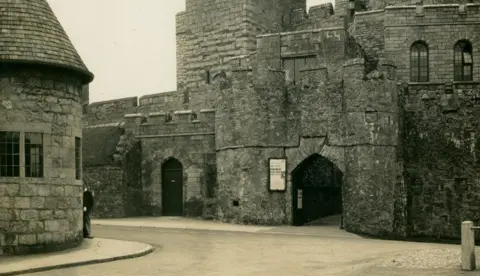 MNH An old photo of the entrance to the castle with the police station on the left and a road running in between them.