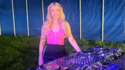 Katie Owen Katie Owen is stood next to turntables inside a tent. She is smiling at the camera, is wearing a pink top and black trousers, and has blonde hair. She's smiling at the camera.