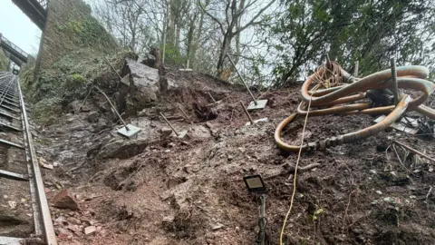 Lynton & Lynmouth Cliff Railway This is a view of part of the cliff, quite close up, looking up from below. It is muddy and rocky. There are a series of pins which have been drilled into the rock, sticking out. The railway track is visible to the left of the image.