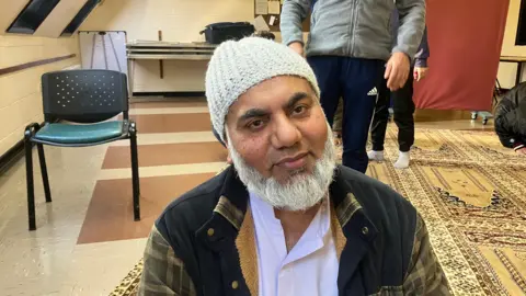 BBC/RICHARD EDWARDS Worshipper Akhtar Zaman - at Selby Friday prayers - is looking slightly to the right of the camera. he has a grey beard and is wearing a white shirt, a white woolly hat and a checked jacket.