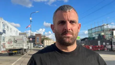 BBC Billy Cooper in a black t-shirt, standing on a pavement. Behind him is a lamp post with a Union flag attached to it.
