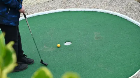A yellow ball sits near a hole on a mini golf course. A child holds a mini golf club nearby. 