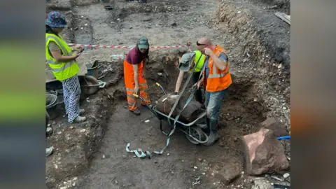 Three volunteers wearing high viz clothing secure the section of column to a wheelbarrow while another is stood next to them taking a picture on her phone. They are in the middle of a dug-up area.