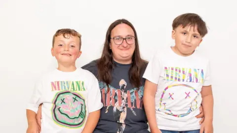 Sarah Haywood Photography Two young boys with brown hair stand either side of a their mother. She has long brown hair, glasses and is wearing a grey Nirvana T-shirt with pink writing and a person with angel wings on it. Both boys are wearing white Nirvana T-shirts with a round smiley face with crosses for eyes. They are all looking at the camera and smiling. 