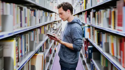 A man with a grey hooded top is standing between full bookshelves and is reading the back of a book.