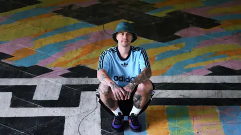 Rikki Hughes Rikki, a man, wearing a blue adidas jersey and black shorts and a round blue hat and purple shoes, squatting on the ground covered with graffiti.