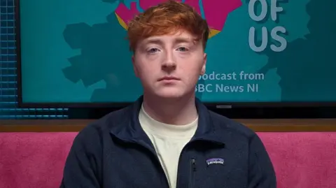 A young man with red hair looks into the camera with a neutral impression on his face. He is wearing a white t-shirt and a blue zip-up fleece jacket. He is sitting in The State of Us podcast studio, which has pink upholstered sofas and a blue walls. Behind his head is a TV screen with The State of Us logo - a pink rendering of the map of Northern Ireland along with the words The State of Us 