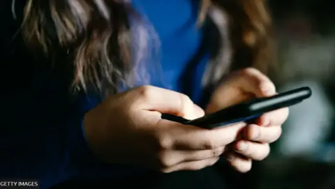 Close up of a young girls hands. She is holding a black smartphone in both hands and it looks like she is reading a message. You can't see her face but you can see she has long brown hair and is wearing a mid blue long sleeved top.