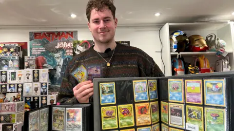 A man with brown hair and a curly fringe is wearing a brown jumper and standing in front of a binder filled with Pokémon cards. He is also holding two cards in his right hand.