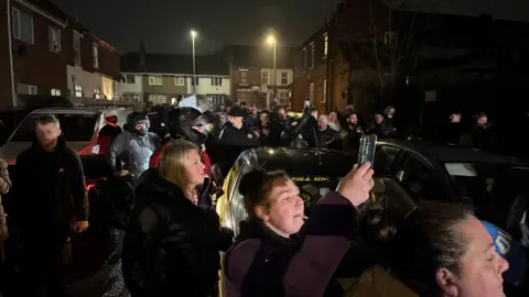 A crowd of people stand in the street in a residential area. Some are holding up their phones.