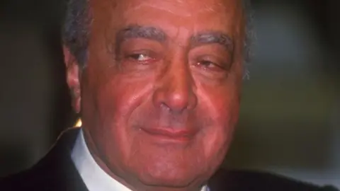 BBC Mohammed Al Fayed com um leve sorriso. Ele está olhando da direita para a esquerda. Suas sobrancelhas são bastante espessas e ele está ficando careca no topo da cabeça. Ele está vestindo camisa branca, gravata escura e paletó preto. A foto foi tirada em 2001.