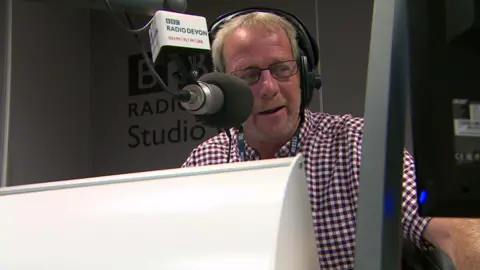 Gordon Sparks in the BBC Radio Devon studio. He is wearing a chequered shirts and headphones. A microphone is pointing towards him.