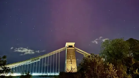 Andrew Cleaver The Northern Lights are visible over the Clifton Suspension Bridge, which is photographed from below at night. The sky is a light purple and green