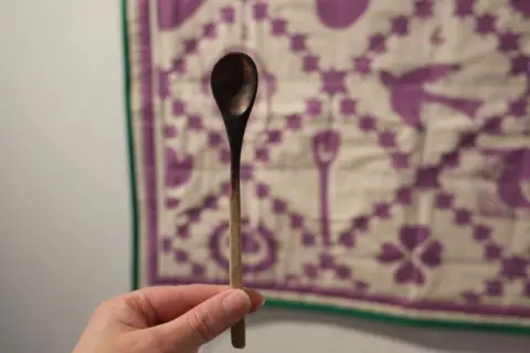 Craftspace A hand can be seen holding up a wooden teaspoon in front of a quilt where an image of it is represented.