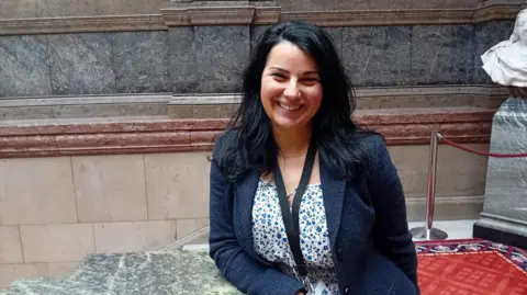 Sheffield Green Party A woman with long black hair is wearing a navy jacket and a white dress with small blue flowers. She is stood on the marble staircase at Sheffield town hall and is smiling