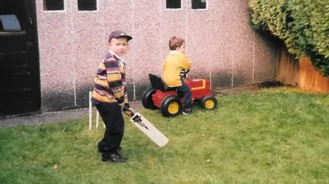 Murray family Matt and Jamie Murray playing in the garden as children. Matt is holding a cricket bat in front of a wicket. Jamie is riding a small red tractor.