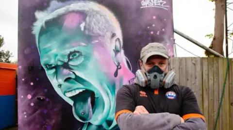 Jaime Bunting Photography David Nash, wearing a face mask, standing in front of his Keith Flint mural - a purple and blue painting showing Keith shouting with his pierced tongue sticking out.