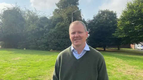 A man in a green sweater and shirt standing in front of some trees