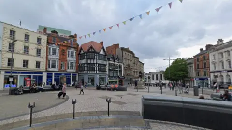 A wide pedestrian square in Wolverhampton city centre, with historic buildings lining the edges, bunting strung overhead and people walking across open paving. Shops and seating areas surround the square under a cloudy sky.