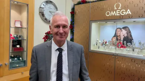 BBC A man wearing a shirt, tie and blazer standing inside a shop. Watches are on display in a cabinet behind him. A clock is on the wall. 