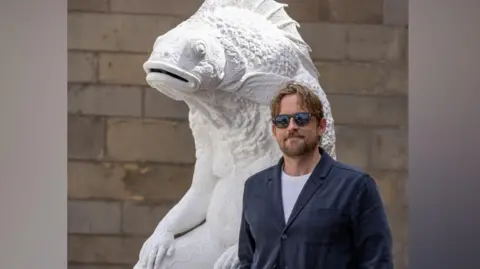 Adam Kendrick Patric Rogers has medium length brown hair and beard and is wearing sunglasses, a white t-shirt and a blue jacket. He is stood next to the sculpture Skalli and is smiling at the camera.