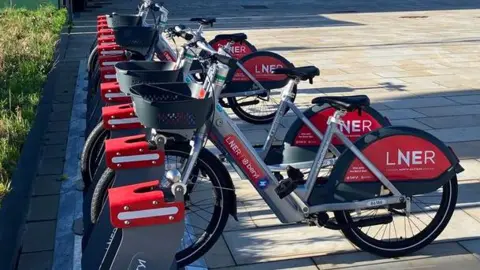 A row of rental e-bikes parked at a docking station in an area of concrete paving slabs, with a small grass park in the distance and buildings behind it