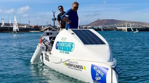 Chloe Foster Tim and Mike stood on their rowing boat. It's white and has various decals on it, along with a solar panel.