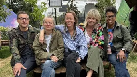 Five people sitting on a wooden chair. Alison Howe has short light-coloured hair and glasses, wearing a green anorak and white T-shirt. Emily Eavis has long brown hair and is wearing a blue and white shirt. There are potted plants behind them.