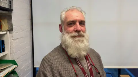 David Birks A man with a long flowing blonde beard looks at the camera while standing in front of a white blind.