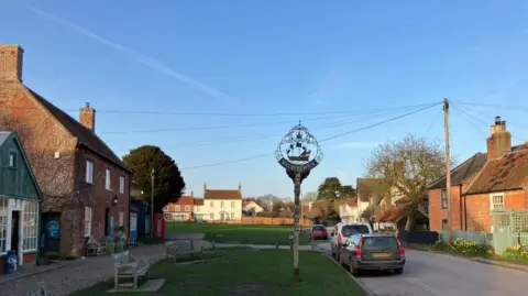 Vikki Irwin/BBC A quiet street in the village. The metal village sign is visible on a patch of grass with another grassy area behind. On either side of the street are old red brick houses. A few cars are parked. A village bench is visible. A white house can be seen beyond.