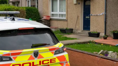 A police car parked outside a ground-floor residential building, with blue-and-white police tape stretched across a dark blue front door, and small garden area.