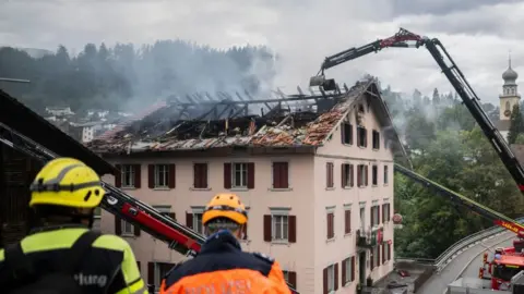 The building seen in the daylight. Smoke rises from the top - most of the roof has been burnt off and charred beams shows. A large crane is removing something from the damaged roof.