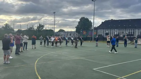 There are around 20 people shown rehearsing their band music. There are those playing the flute and drummers also. The picture is a wide shot of the band members standing on a football pitch. 