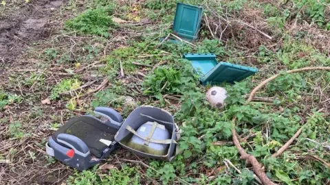 Torridge District Council A child's car seat, a foot ball and a green plastic container have been dumped at a beauty spot. The waste items are scattered across the green foliage. Mud, sticks and stinging nettles are on the ground. 