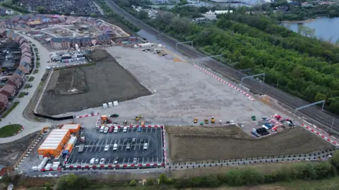 Ant Saddington/BBC An aerial view of the construction of a railway station. There is a railway line with an open space next to it along with a small car park. On the right of the railway line there are trees. There are houses on the left and top left of the picture.