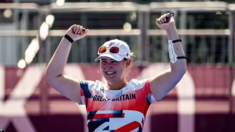 Getty Images Phoebe Paterson Pine is wearing a red, white and blue t-shirt that says "Great Britain". She has her arms in the air, celebrating. She is smiling and wearing a white cap, with white sunglasses resting on it