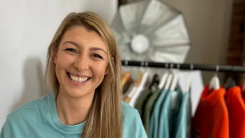 BBC/Helen Richardson Claire Goodliff is pictured wearing one of the t-shirts she designed. Behind her there is a rail of her clothes. She has long brown hair with blonde highlights. 
