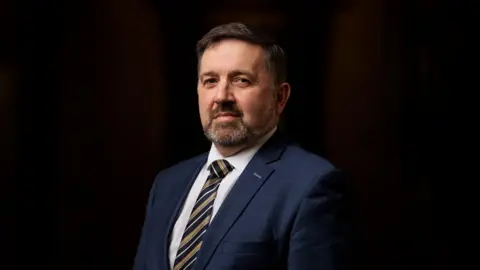 PA Robin Swann wears a white shit and a blue blazer. He has a grey beard and greying hair. Behind him is a black backdrop.  