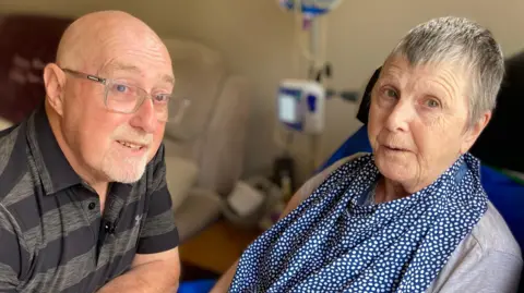 Steve Duckett, who is 72, bald with glasses and a goatee beard sits with his wife Sandy, aged 72. She has Huntington's disease. She sits in an adapted chair and has a material bib around her neck