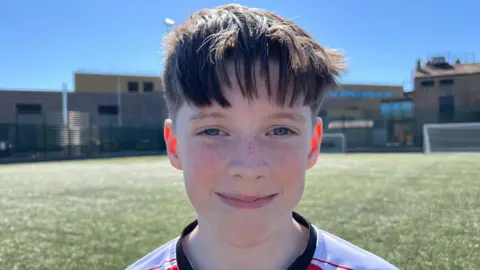 A boy is wearing a white and red football top. He has brown hair. He is smiling.