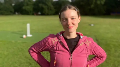  A woman wearing a pink zip up top. She is stood in a field with cricket stumps in the background.